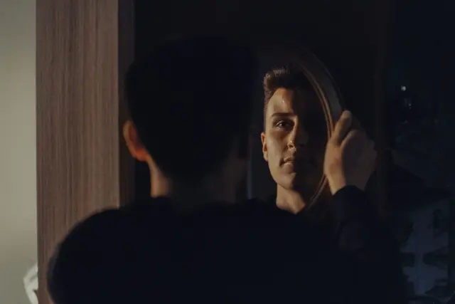 A person standing before a mirror in a softly lit bedroom, seen from behind.
