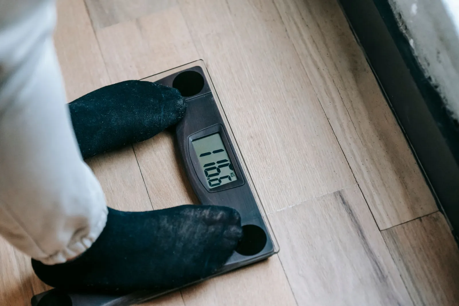 A person in socks standing on a digital bathroom scale on a wood floor, checking their weight.