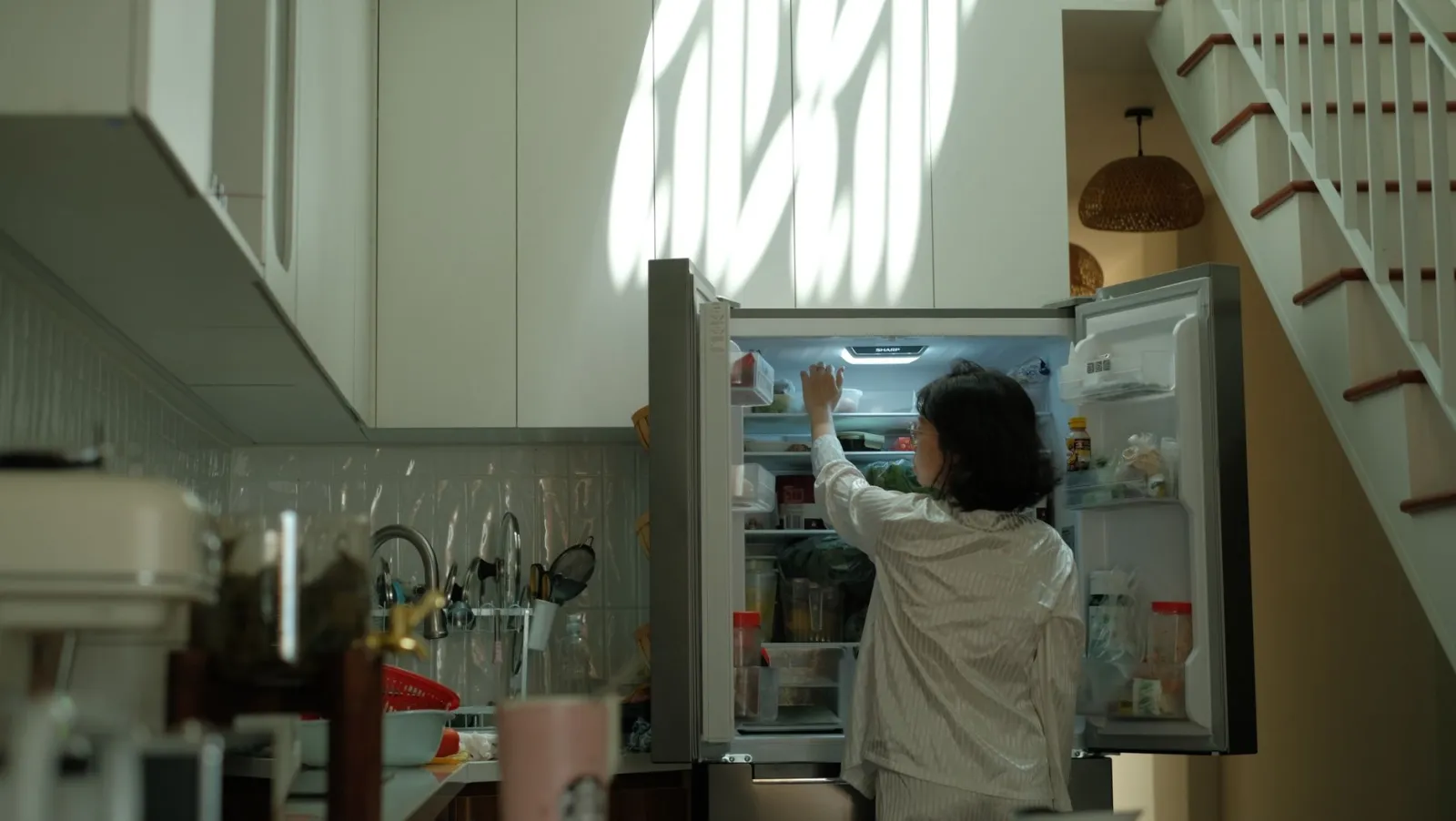 A woman in pajamas checks the refrigerator in a warmly lit cozy kitchen.