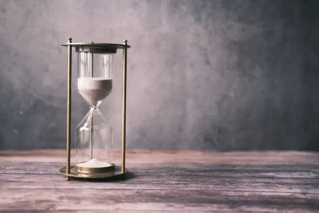 A classic hourglass with sand on a rustic wooden table against a soft grey background.