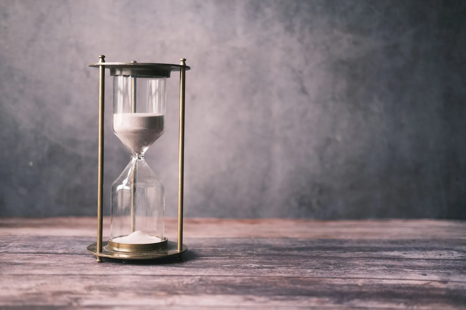 A classic hourglass with sand on a rustic wooden table against a soft grey background.