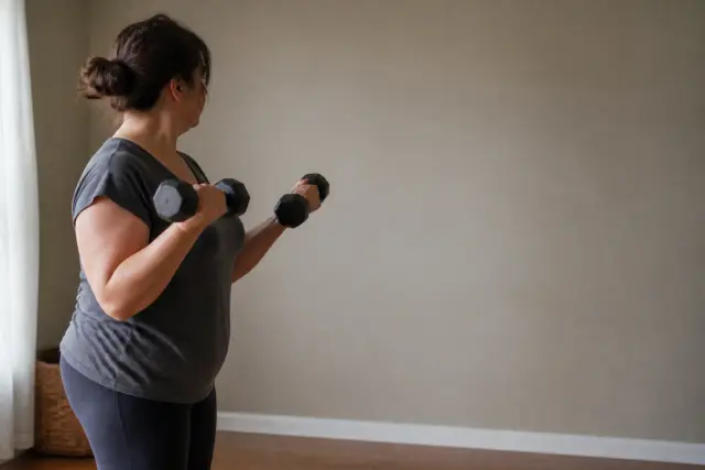 A middle-aged woman lifting a pair of dumbbells in a plain room in flat daylight.