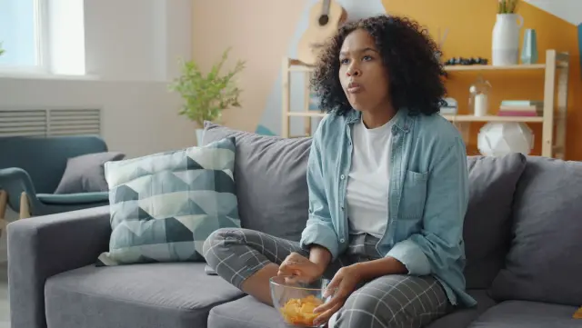 A woman sitting on a couch with a bowl of snacks beside her, watching a screen with a flat expression.
