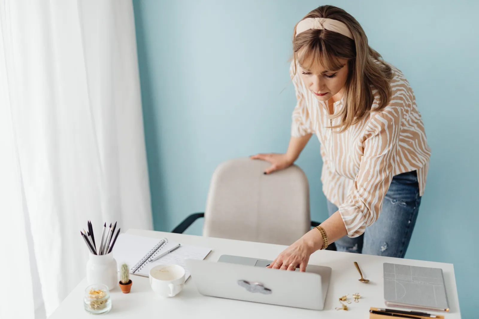 A woman standing at a desk using a laptop beside a notepad and coffee cup.