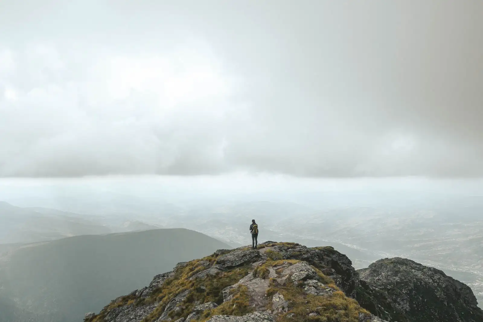 A lone hiker standing still on a mountain peak under a wide cloudy sky, at the top of the climb.