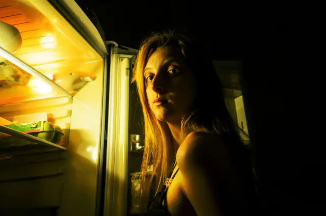 A person standing in a dim kitchen at night, looking thoughtfully into an open refrigerator.