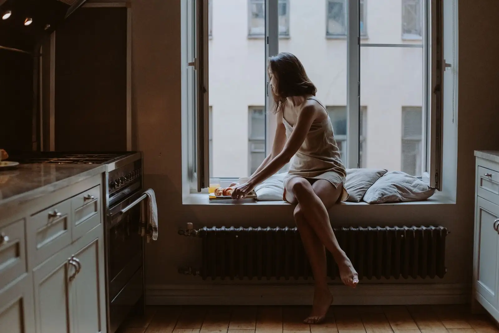 A woman sitting by a bright kitchen window with a small tray of food, eating calmly and without hurry.