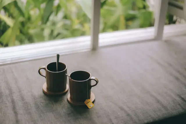 Two warm cups on a table by a large window with greenery outside.